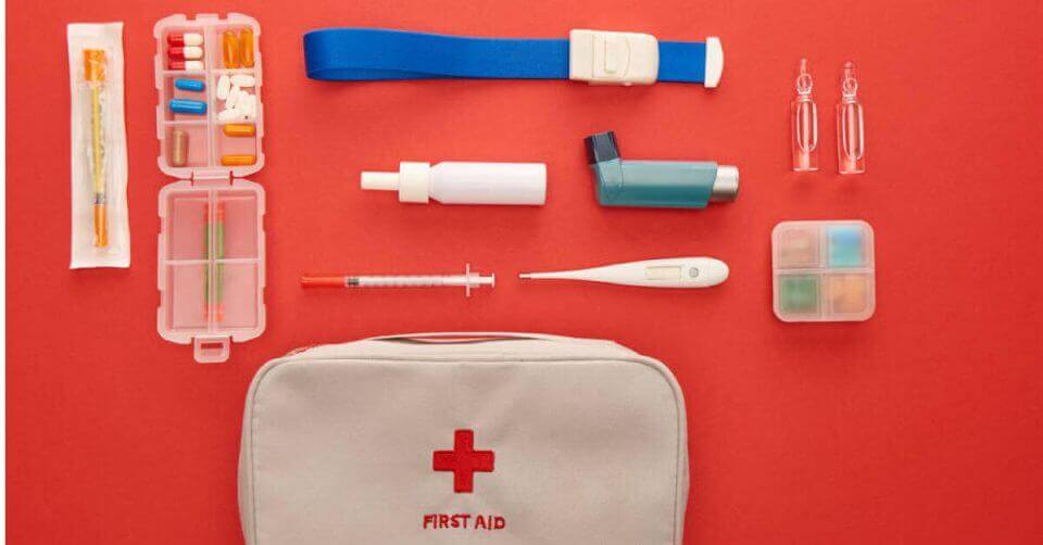 First aid kit with inhaler, thermometer, tourniquet, syringe, and medicine capsules on a red background.