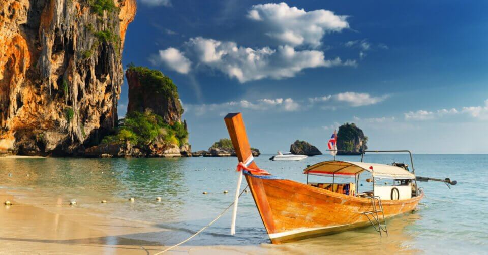 Wooden boat on a sandy beach with turquoise water, limestone cliffs, and a clear blue sky with scattered clouds.