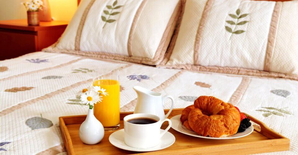 Breakfast tray with coffee, orange juice, a croissant, and flowers on a bed with floral-patterned pillows and quilt.