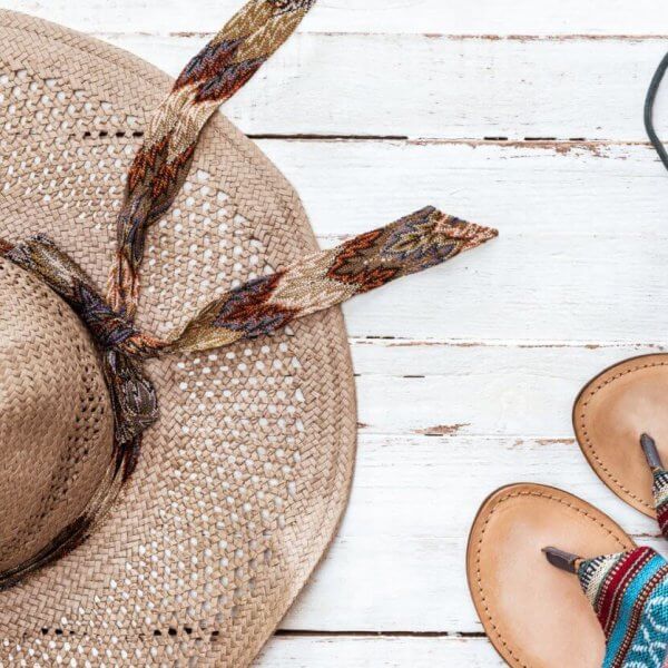 A straw hat with a patterned ribbon, a pair of leather sandals featuring colorful woven straps, and black earphones are artistically arranged on a white wooden surface. Evoking a casual, summery vibe perfect for Philippine holidays in 2024.