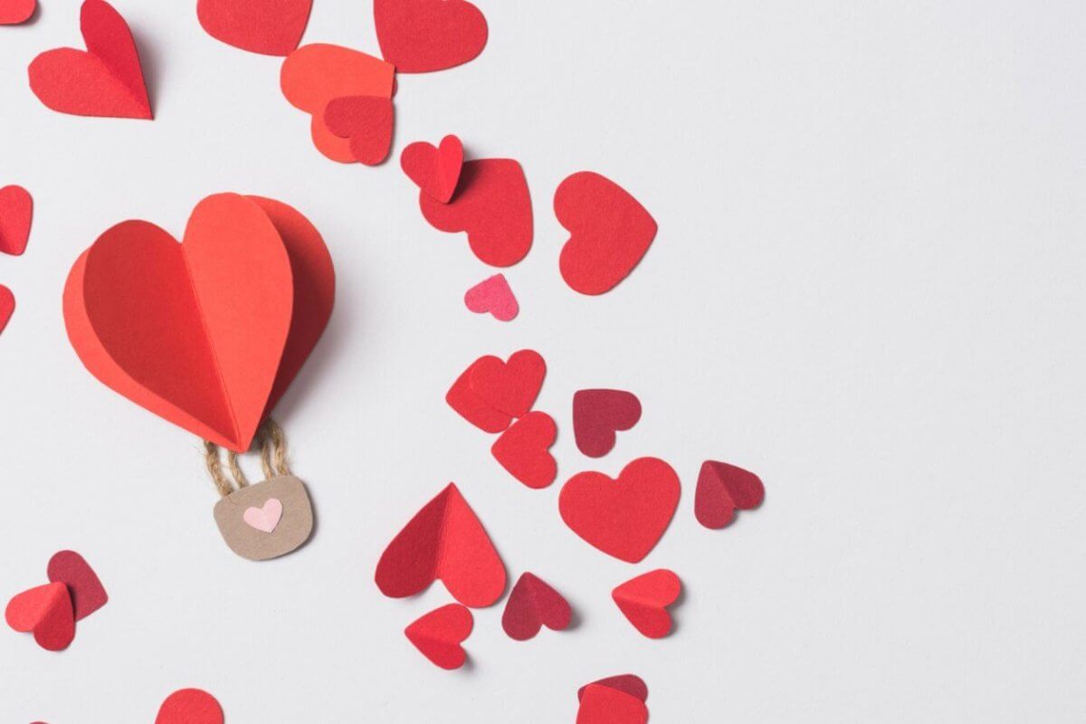 A paper hot air balloon with a red heart floats among scattered red and pink heart-shaped cutouts on a white background. Perfect for 2023 Valentine Gift Ideas, the whimsical scene features a brown basket with two strings, capturing love's playful essence.