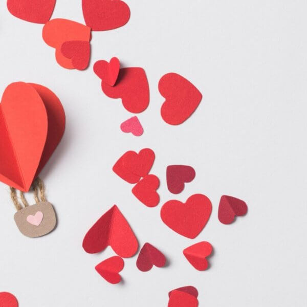 A paper hot air balloon with a red heart floats among scattered red and pink heart-shaped cutouts on a white background. Perfect for 2023 Valentine Gift Ideas, the whimsical scene features a brown basket with two strings, capturing love's playful essence.
