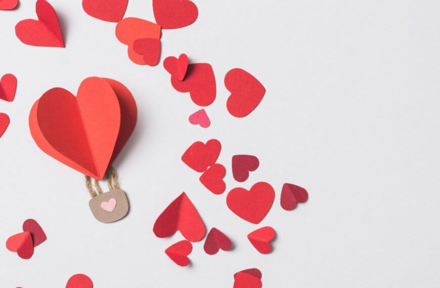 A paper hot air balloon with a red heart floats among scattered red and pink heart-shaped cutouts on a white background. Perfect for 2023 Valentine Gift Ideas, the whimsical scene features a brown basket with two strings, capturing love's playful essence.