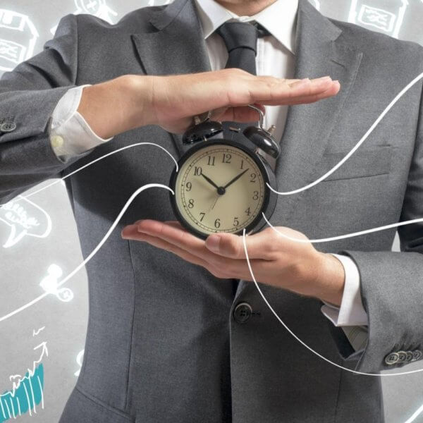 A man in a gray suit holds a traditional alarm clock with both hands against a background filled with various colorful doodles, including charts, graphs, a light bulb, a laptop, and other business-related symbols. The clock's hands point to 10:10. Lines connect the clock to the doodles, symbolizing 2023 business ideas.