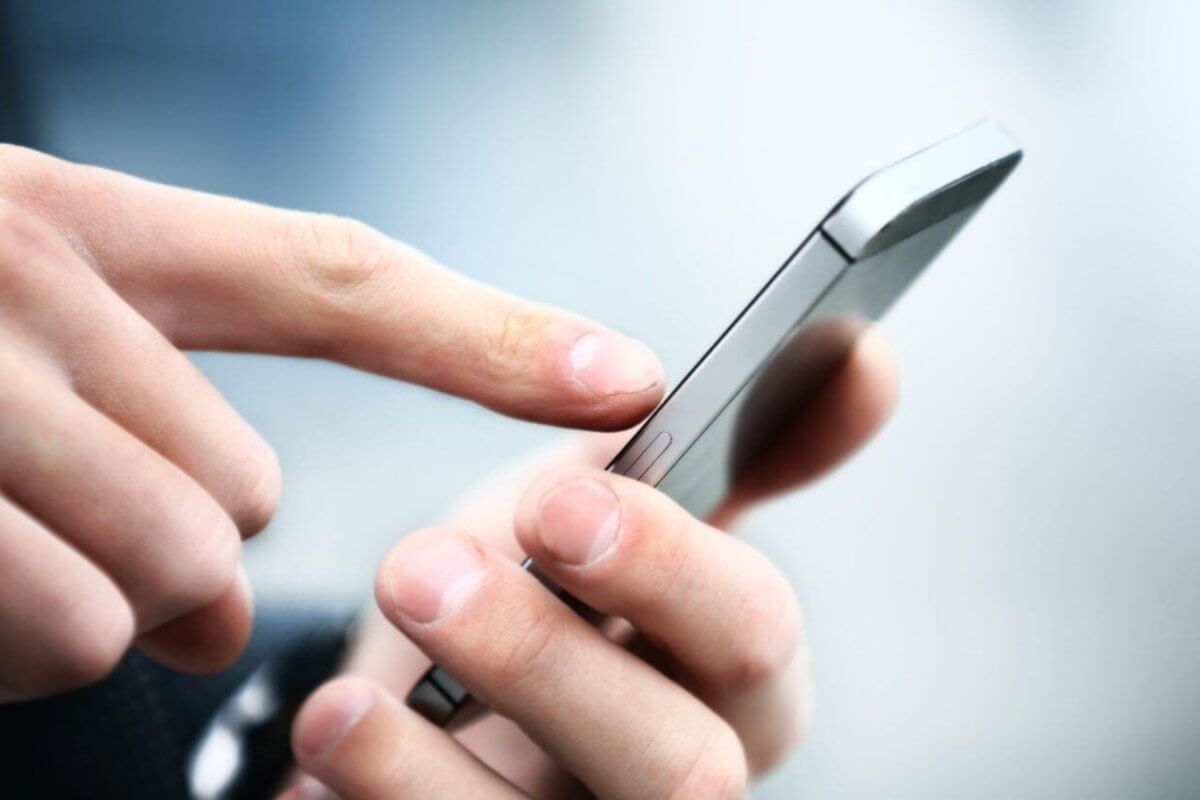 Close-up image of a person’s hands holding a sleek, new phone. The person is using their right index finger to touch the screen. The background is blurred with a mix of light and dark blue tones, focusing attention on the device and hands. The smartphone has a metallic edge, highlighting its modern design.