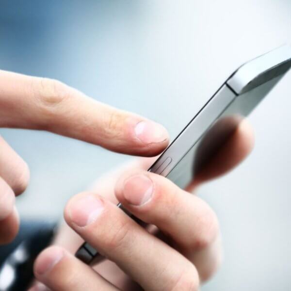 Close-up image of a person’s hands holding a sleek, new phone. The person is using their right index finger to touch the screen. The background is blurred with a mix of light and dark blue tones, focusing attention on the device and hands. The smartphone has a metallic edge, highlighting its modern design.