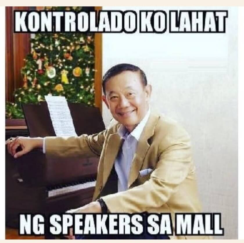 A man in a beige suit and blue shirt sits at a piano with a sheet of music, smiling at the camera like he's part of the latest Christmas memes. Behind him is a decorated Christmas tree. Text in bold above reads "KONTROLADO KO LAHAT," and below, "NG SPEAKERS SA MALL.