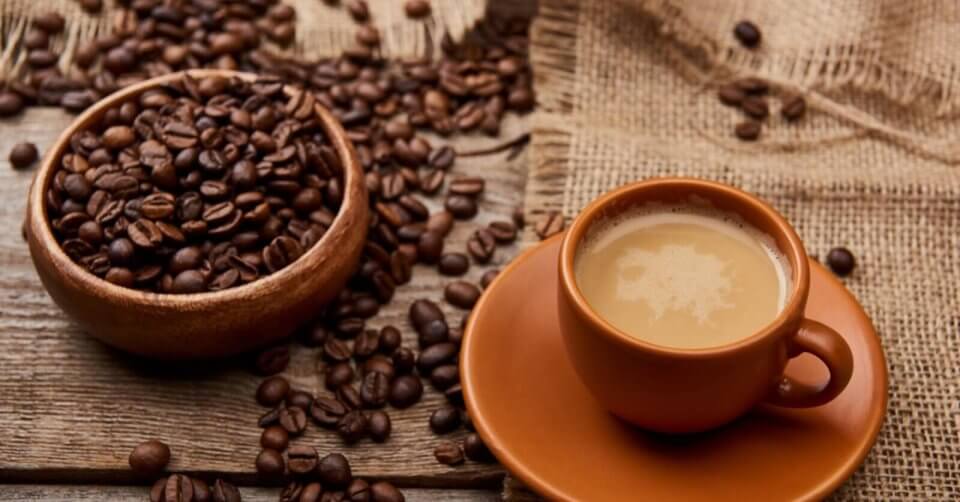 A wooden bowl overflowing with Manila coffee beans sits next to an espresso cup filled with coffee on a saucer, all placed on a rustic wooden table partially covered by a burlap cloth.