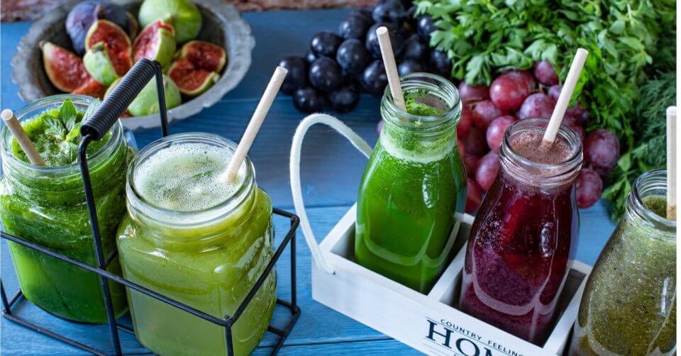 Four glass jars filled with colorful smoothies—green, dark purple, and another green variant—sit in a white wooden tray. Each jar has a straw and is encircled by fresh figs, dark grapes, and leafy greens atop a blue wooden table, capturing the vibrant vibe of Metro Manila juice bars like Juice Up.