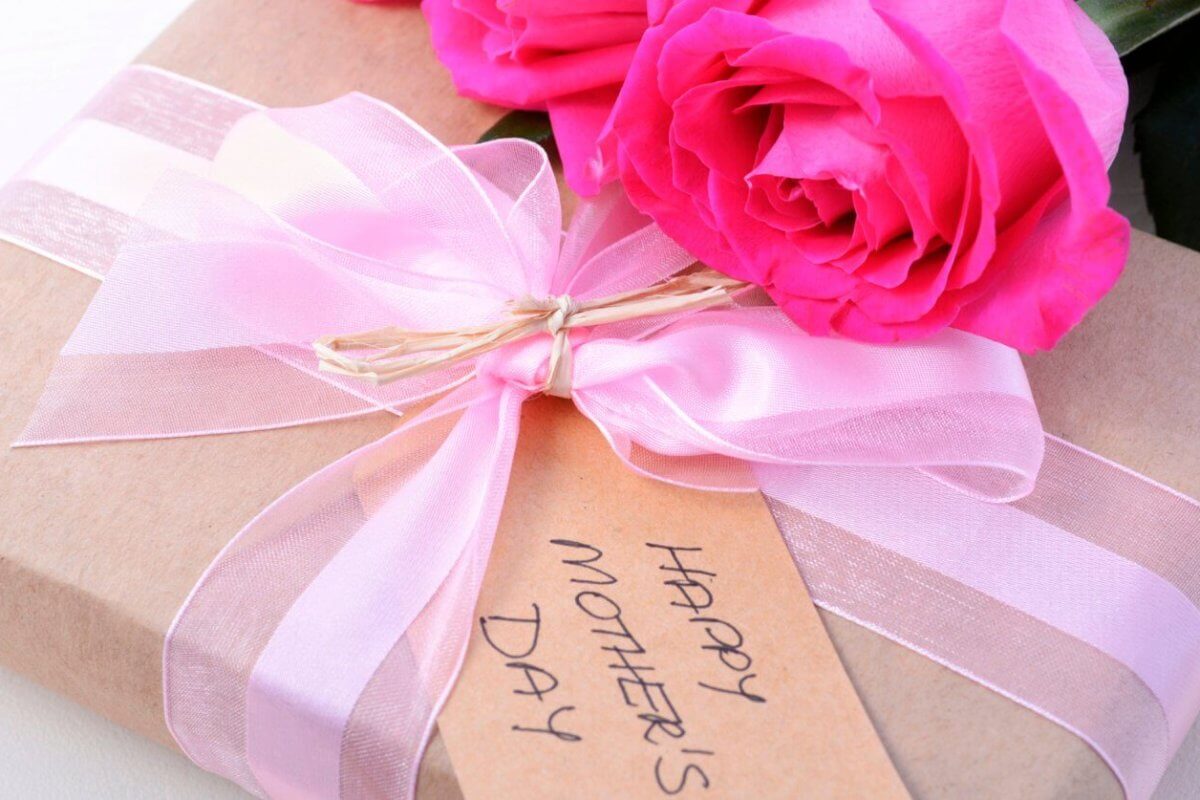 A close-up of a gift wrapped in brown paper tied with a pink ribbon. A tag reading "Happy Mother's Day" is attached to the ribbon, and two bright pink roses rest beside the gift, perfect for celebrating Mother's Day.
