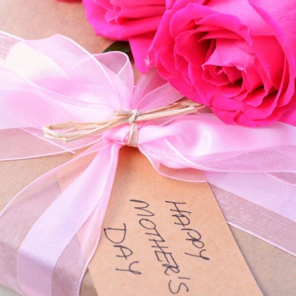 A close-up of a gift wrapped in brown paper tied with a pink ribbon. A tag reading "Happy Mother's Day" is attached to the ribbon, and two bright pink roses rest beside the gift, perfect for celebrating Mother's Day.