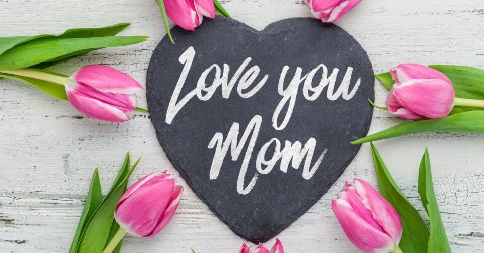 A slate heart with "Love you Mom" written in white, surrounded by pink tulips on a rustic white wooden surface. The arrangement and flowers create a festive, loving atmosphere, perfect to celebrate Mother's Day.