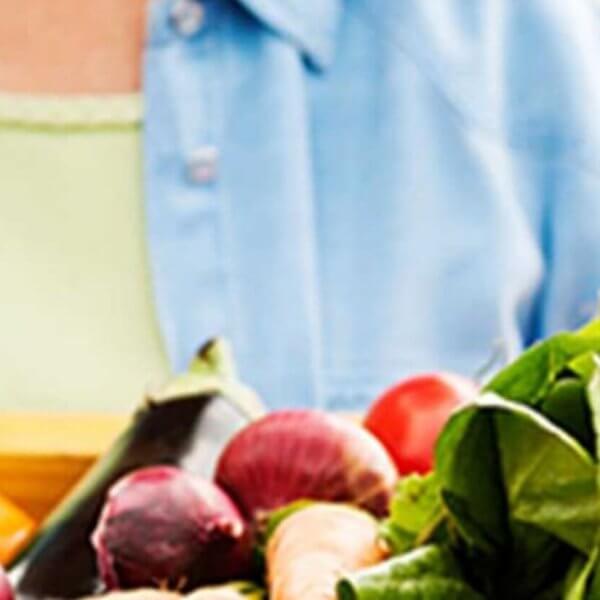 A person holding a wooden basket full of vibrant fresh vegetables including red apples, green lettuce, and purple beets, all sourced from organic hotspots, with a focus on healthy eating. The person wears a light green shirt and blue jacket.