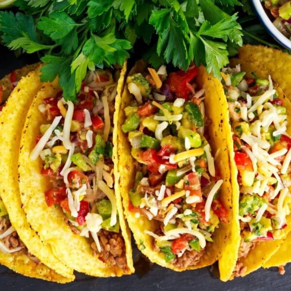 Three crunchy tacos filled with seasoned ground beef, diced tomatoes, chopped onions, shredded cheese, and garnished with cilantro. The tacos are placed on a dark surface surrounded by halved avocados, red chili peppers, a bowl of salsa, and a bunch of cherry tomatoes. Discover the best taco places inspired by Taco Places Metro Manila.
