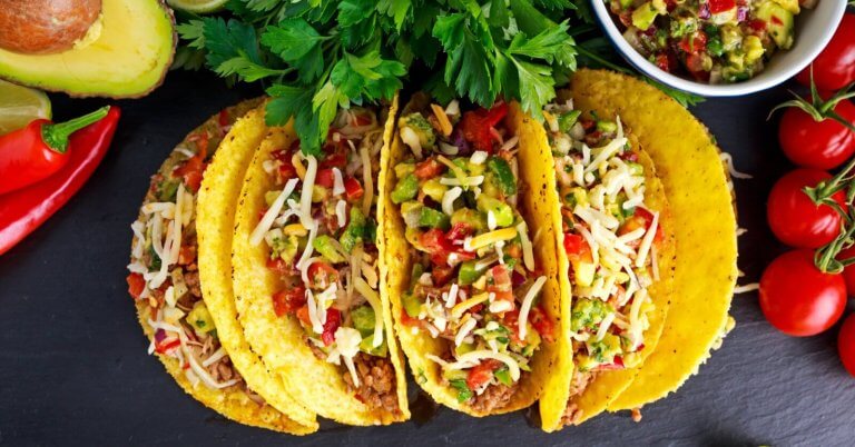Three crunchy tacos filled with seasoned ground beef, diced tomatoes, chopped onions, shredded cheese, and garnished with cilantro. The tacos are placed on a dark surface surrounded by halved avocados, red chili peppers, a bowl of salsa, and a bunch of cherry tomatoes. Discover the best taco places inspired by Taco Places Metro Manila.