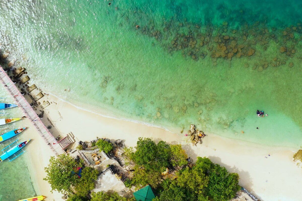 Aerial view of Alaminos City beach with turquoise waters and a thin line of white sand. A pier extends from the shore on the left, near colorful boats docked. Swimmers are visible in the clear water, with patches of rocks observable underwater. Lush green trees line the edge, perfect for post-swim visits to must-try restaurants nearby.