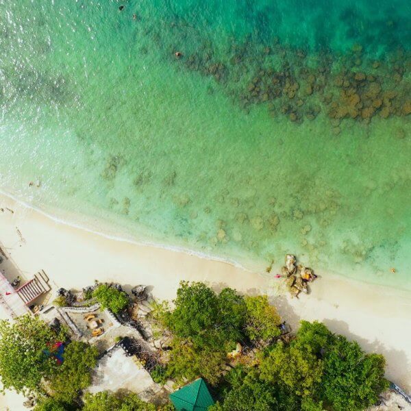 Aerial view of Alaminos City beach with turquoise waters and a thin line of white sand. A pier extends from the shore on the left, near colorful boats docked. Swimmers are visible in the clear water, with patches of rocks observable underwater. Lush green trees line the edge, perfect for post-swim visits to must-try restaurants nearby.