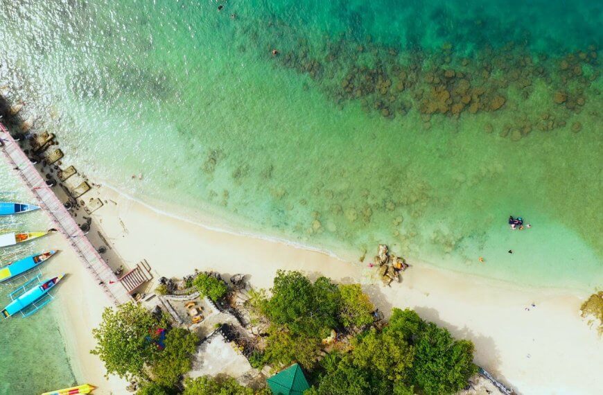 Aerial view of Alaminos City beach with turquoise waters and a thin line of white sand. A pier extends from the shore on the left, near colorful boats docked. Swimmers are visible in the clear water, with patches of rocks observable underwater. Lush green trees line the edge, perfect for post-swim visits to must-try restaurants nearby.