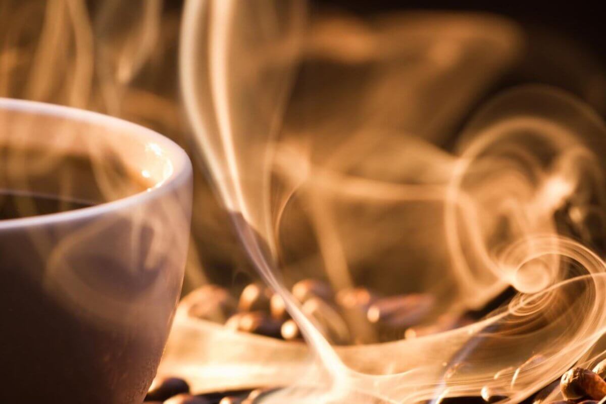 A white cup filled with steaming hot coffee sits on a surface scattered with coffee beans at a Manila 24/7 coffee shop. The steam rises in delicate, swirling patterns, creating an artistic effect in the warm, ambient lighting. The focus is close-up, capturing the texture of the beans and the ethereal quality of the steam.