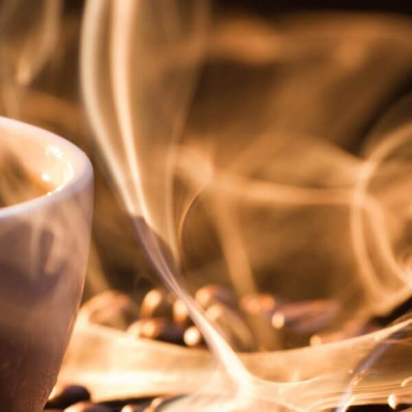 A white cup filled with steaming hot coffee sits on a surface scattered with coffee beans at a Manila 24/7 coffee shop. The steam rises in delicate, swirling patterns, creating an artistic effect in the warm, ambient lighting. The focus is close-up, capturing the texture of the beans and the ethereal quality of the steam.