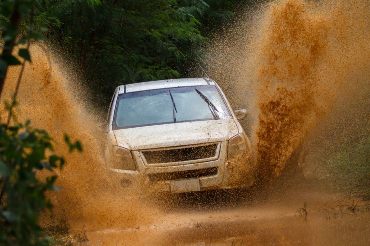 A white SUV drives through a muddy terrain, splashing mud high into the air. The vehicle is surrounded by lush green foliage, showcasing the adventure and rugged environment. The dynamic shot captures the energy of visiting Metro Manila's best 4x4 offroad shops and experiencing an exhilarating off-road journey.