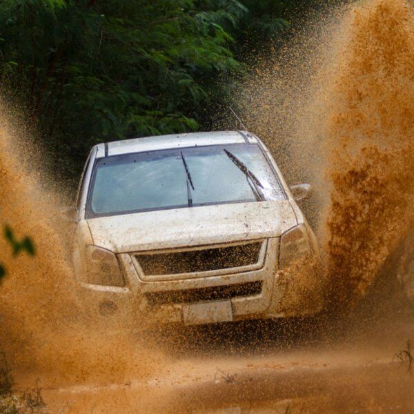 A white SUV drives through a muddy terrain, splashing mud high into the air. The vehicle is surrounded by lush green foliage, showcasing the adventure and rugged environment. The dynamic shot captures the energy of visiting Metro Manila's best 4x4 offroad shops and experiencing an exhilarating off-road journey.