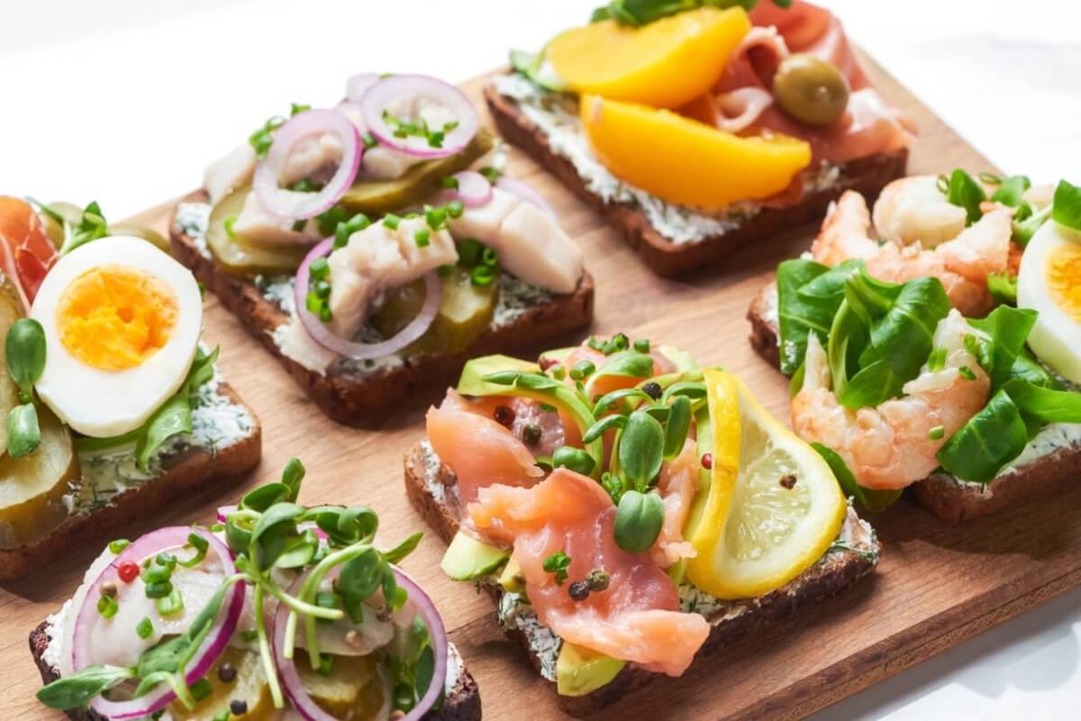 A wooden board topped with six open-faced sandwiches, a must-try at Sulit Restaurants in El Nido, featuring colorful toppings like sliced boiled eggs, pickles, red onions, various greens, salmon, shrimp, lemon slices, peach slices, and olives on dark bread. The sandwiches are neatly arranged in two rows of three.