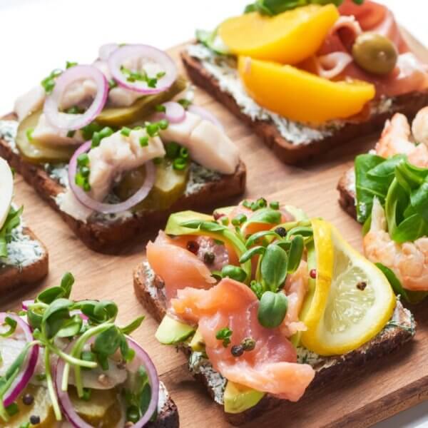 A wooden board topped with six open-faced sandwiches, a must-try at Sulit Restaurants in El Nido, featuring colorful toppings like sliced boiled eggs, pickles, red onions, various greens, salmon, shrimp, lemon slices, peach slices, and olives on dark bread. The sandwiches are neatly arranged in two rows of three.