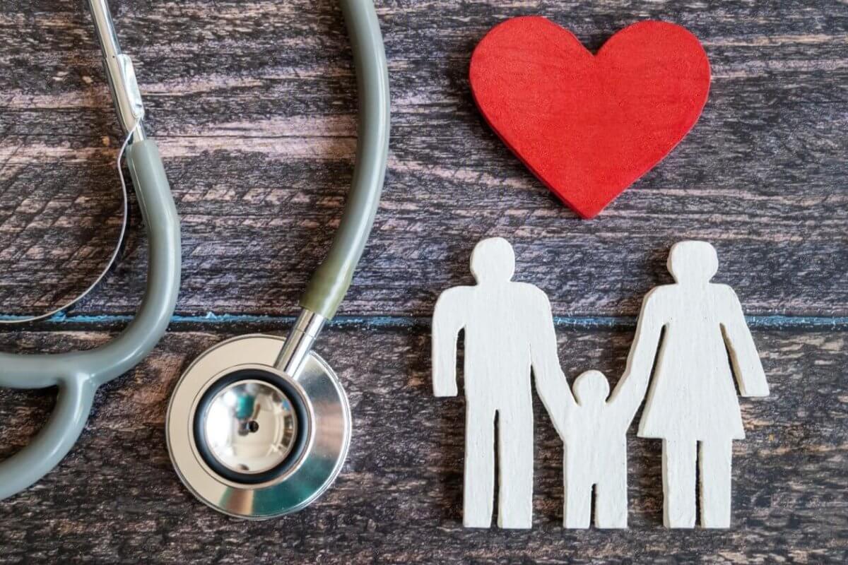 A stethoscope is positioned on the left side of the image, with its ear tubes and chest piece visible. A red wooden heart lies to the right of the stethoscope. Next to the heart, there's a white wooden cutout depicting a family of three holding hands. This scene evokes Hi-Precision Diagnostics centers in Metro Manila.