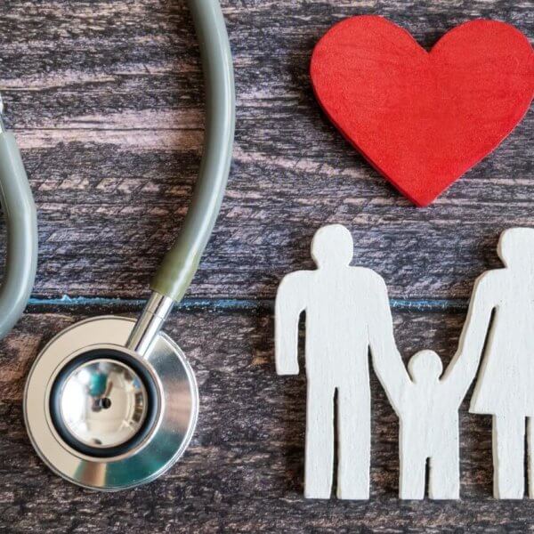 A stethoscope is positioned on the left side of the image, with its ear tubes and chest piece visible. A red wooden heart lies to the right of the stethoscope. Next to the heart, there's a white wooden cutout depicting a family of three holding hands. This scene evokes Hi-Precision Diagnostics centers in Metro Manila.