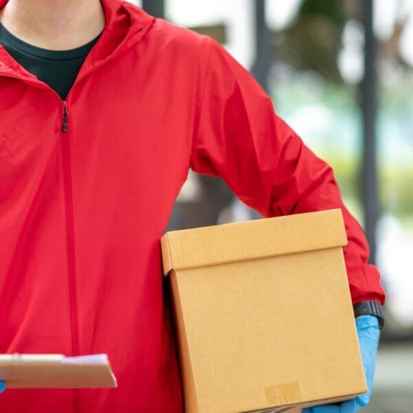 A person wearing a red jacket and blue gloves holds a brown cardboard box in one hand and a clipboard in the other. The background is slightly blurred, showing an indoor setting with large glass windows and greenery outside, possibly indicating J&T Drop-Off Locations within Metro Manila, emphasizing convenience.