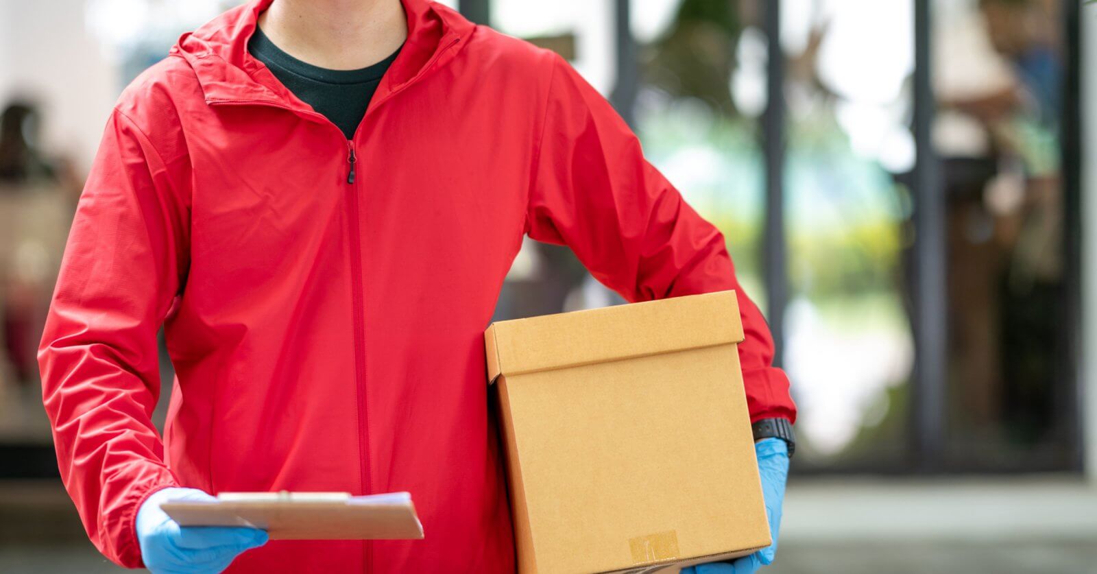 A person wearing a red jacket and blue gloves holds a brown cardboard box in one hand and a clipboard in the other. The background is slightly blurred, showing an indoor setting with large glass windows and greenery outside, possibly indicating J&T Drop-Off Locations within Metro Manila, emphasizing convenience.