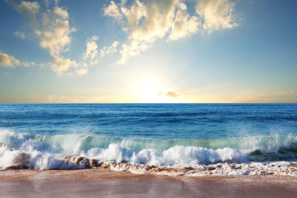 A scenic view of a Zambales beach at sunrise with gentle waves lapping the shore. The golden sunlight illuminates the sky, casting a warm glow on scattered clouds. The calm ocean stretches to the horizon, blending into the sky where the sun peeks just above the water's edge—truly one of the best Zambales resorts.