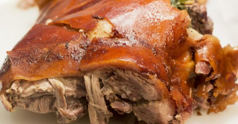 A close-up of a roasted pork dish from one of the best restaurants in Alabang, showcasing crispy, golden-brown skin with some tender, juicy shredded meat visible underneath. The meat appears well-cooked and succulent, with a slightly charred, flavorful exterior. The plain white background highlights the dish's texture.
