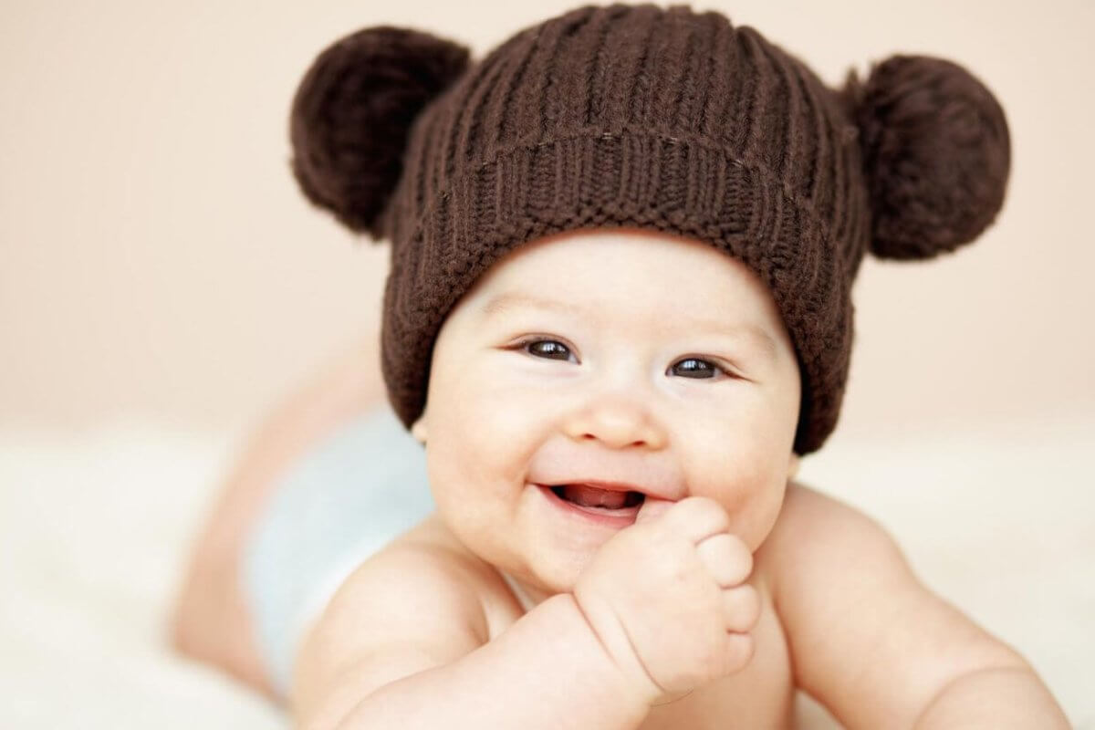 A smiling baby lies on their stomach, wearing a brown knitted hat with two pom-poms resembling bear ears. The baby’s hand is near their mouth, and they have a light-colored diaper on. The background is soft beige, adding to the warm and cozy feel of the image—perfect for showcasing baby essentials from the best baby stores.