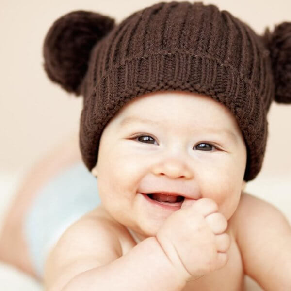 A smiling baby lies on their stomach, wearing a brown knitted hat with two pom-poms resembling bear ears. The baby’s hand is near their mouth, and they have a light-colored diaper on. The background is soft beige, adding to the warm and cozy feel of the image—perfect for showcasing baby essentials from the best baby stores.