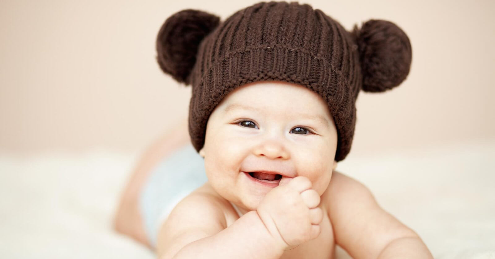 A smiling baby lies on their stomach, wearing a brown knitted hat with two pom-poms resembling bear ears. The baby’s hand is near their mouth, and they have a light-colored diaper on. The background is soft beige, adding to the warm and cozy feel of the image—perfect for showcasing baby essentials from the best baby stores.