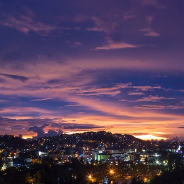 A panoramic view of a city at dusk with a vibrant, multicolored sky. The horizon showcases purple, pink, and orange hues as the sun sets. City lights flicker below, illuminating buildings and streets. Silhouetted clouds and hills add depth to the scene. The atmosphere is calm and picturesque—the perfect backdrop for Baguio Vacation Rentals or Budget Stays in Baguio Homes.
