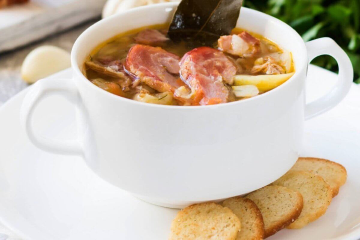 A white bowl filled with a hearty soup reminiscent of Tagaytay's famous Bulalo, containing chunks of meat, bone and vegetables, garnished with a leaf, is set on a white plate with three slices of toasted bread on the side. The background features ingredients like garlic and fresh greens, hinting at a rustic, homemade meal often celebrated in restaurants.