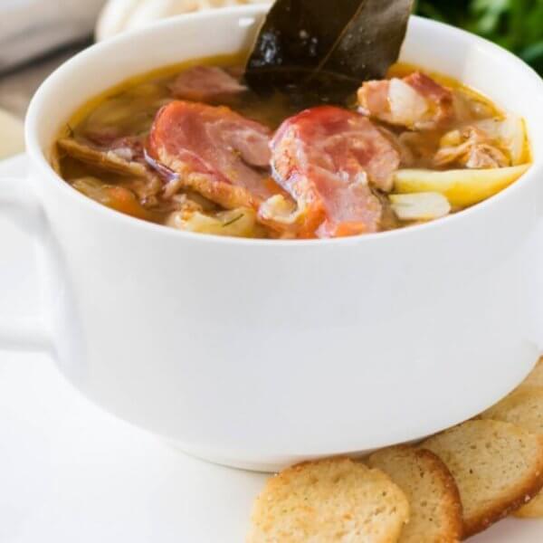 A white bowl filled with a hearty soup reminiscent of Tagaytay's famous Bulalo, containing chunks of meat, bone and vegetables, garnished with a leaf, is set on a white plate with three slices of toasted bread on the side. The background features ingredients like garlic and fresh greens, hinting at a rustic, homemade meal often celebrated in restaurants.