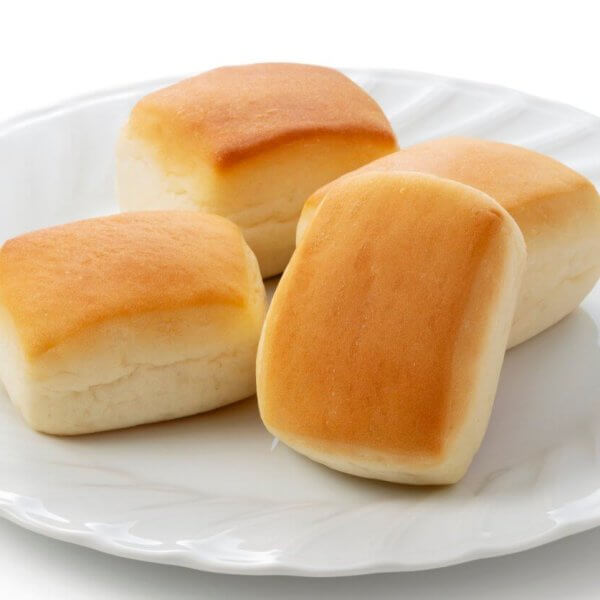 A white plate holds four golden-brown dinner rolls, each with a soft, fluffy texture. The rolls are slightly rectangular with rounded edges, and their tops are a light golden color that gradually lightens toward the bottom. Reminiscent of the best hot pandesal from Metro Manila's must-try bakeries, this plate features subtle decorative ridges around the rim.