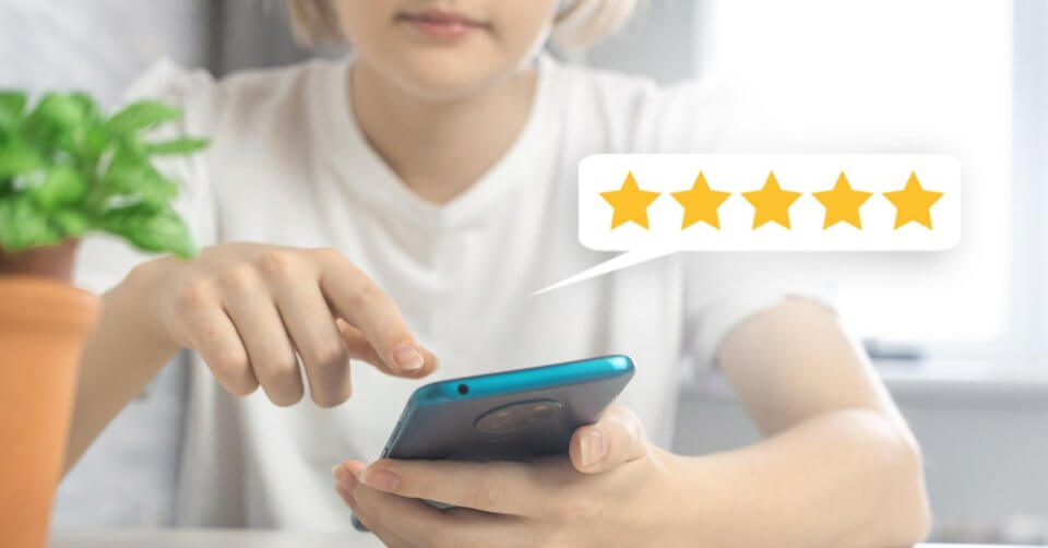 A person in a white shirt uses a smartphone at a table, their finger gestures swiftly on the screen. A speech bubble above showcases a five-star rating, hinting at tips from a phone buying guide. A blurred plant in the foreground adds to the softly lit, peaceful ambiance.