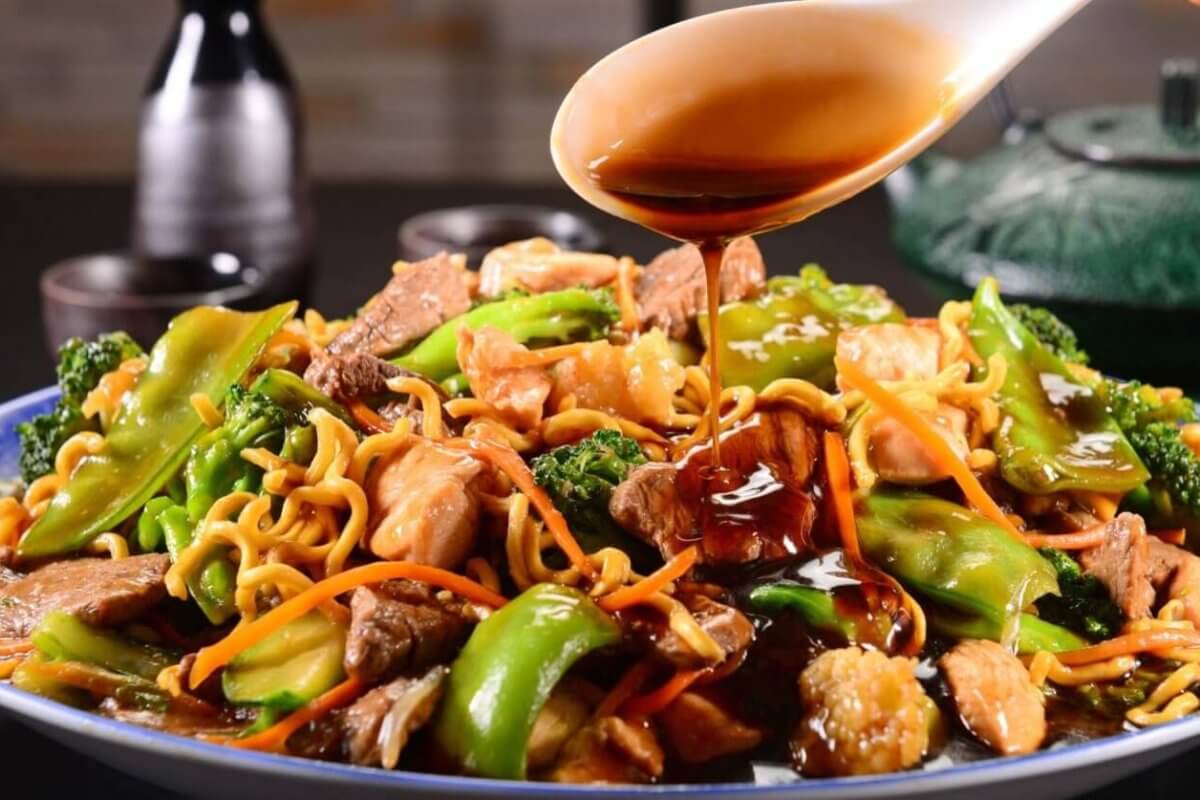 A close-up of a plate of stir-fried noodles loaded with vegetables like broccoli, bell peppers, and carrots, alongside sliced meat and shrimp. A hand is pouring a dark, glossy sauce from a spoon over the dish. In the background, two small bottles and a teapot are visible on a blurred dark surface—a scene reminiscent of the best Binondo restaurants.