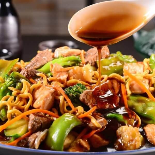 A close-up of a plate of stir-fried noodles loaded with vegetables like broccoli, bell peppers, and carrots, alongside sliced meat and shrimp. A hand is pouring a dark, glossy sauce from a spoon over the dish. In the background, two small bottles and a teapot are visible on a blurred dark surface—a scene reminiscent of the best Binondo restaurants.