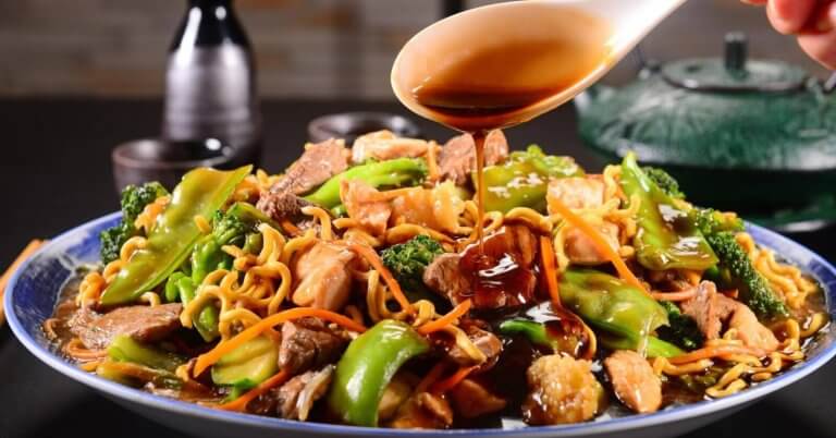 A close-up of a plate of stir-fried noodles loaded with vegetables like broccoli, bell peppers, and carrots, alongside sliced meat and shrimp. A hand is pouring a dark, glossy sauce from a spoon over the dish. In the background, two small bottles and a teapot are visible on a blurred dark surface—a scene reminiscent of the best Binondo restaurants.