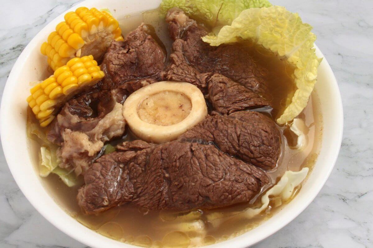 A bowl of hearty Bulalo soup featuring large chunks of tender beef, a central beef bone with marrow, corn on the cob pieces, and napa cabbage leaves in a clear broth. This Filipino favorite, popular in Metro Manila hotspots, is served in a white bowl set on a marble surface.
