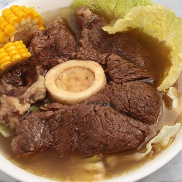 A bowl of hearty Bulalo soup featuring large chunks of tender beef, a central beef bone with marrow, corn on the cob pieces, and napa cabbage leaves in a clear broth. This Filipino favorite, popular in Metro Manila hotspots, is served in a white bowl set on a marble surface.