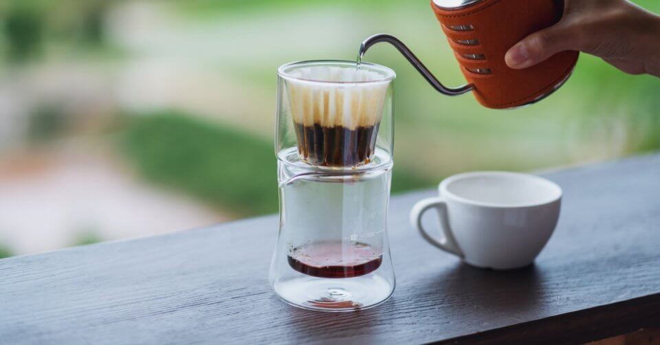 A hand pours hot water from a kettle over a coffee dripper sitting on a double-walled glass with brewed coffee inside, creating the perfect blend for relaxing at one of Tanay's serene coffee spots. A white cup rests beside it on a wooden surface against a backdrop of blurred, green outdoor landscape.