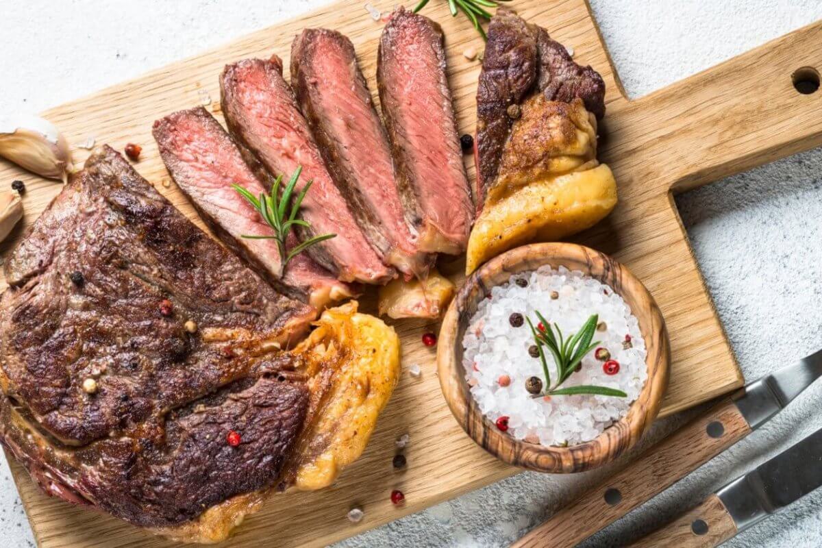 Grilled steak sliced into pieces on a wooden cutting board with a handle. Black and red pepper sprinkled on the steak. A wooden bowl filled with coarse salt and peppercorns sits next to the steak. Garnished with rosemary sprigs, garlic cloves, and accompanied by a fork and knife on the side—an experience at one of Eastwood City's must-visit restaurants.