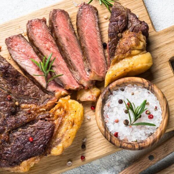 Grilled steak sliced into pieces on a wooden cutting board with a handle. Black and red pepper sprinkled on the steak. A wooden bowl filled with coarse salt and peppercorns sits next to the steak. Garnished with rosemary sprigs, garlic cloves, and accompanied by a fork and knife on the side—an experience at one of Eastwood City's must-visit restaurants.
